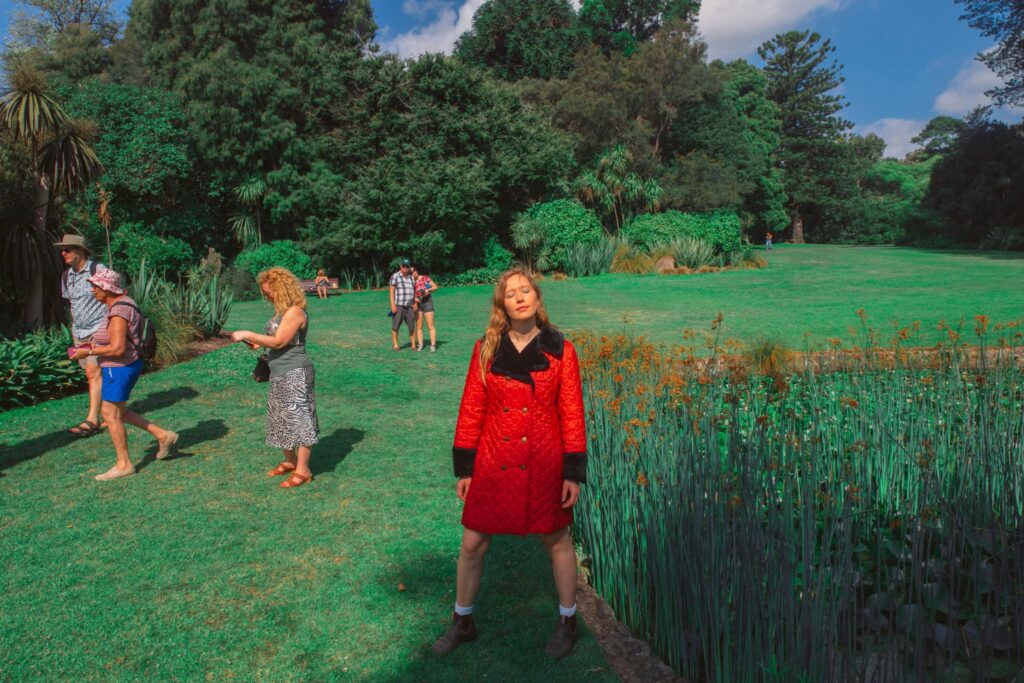 A photo of Julia Jacklin wearing a bright red coat with black lapels and cuffs standing with her eyes closed in the middle of a lush green park. There are some people in the background not paying any attention to her.