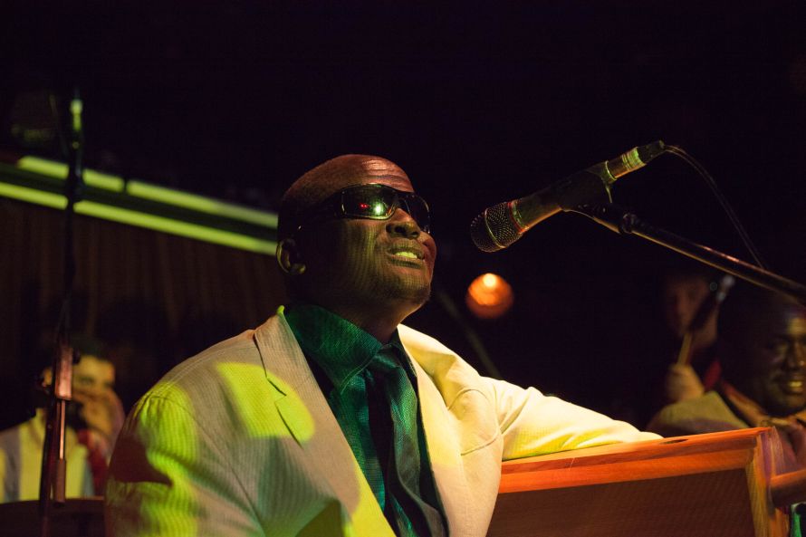 ID: A close up photo of Gordon Koang singing into a microphone on stage. He is wearing a white jacket with an emerald green shirt underneath. He has sunglasses on.