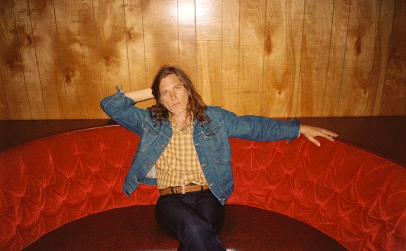 A photo of Eric D Johnson sitting on a red curved sofa. He is wearing a yellow shirt and blue denim jacket and is looking at the camera.
