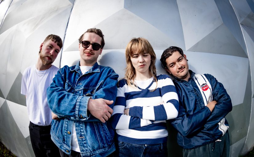 A photo of the band standing together in front of an angulated white background. They are wearing various shades of blue and there is a slighty fisheye distortion to the photo.